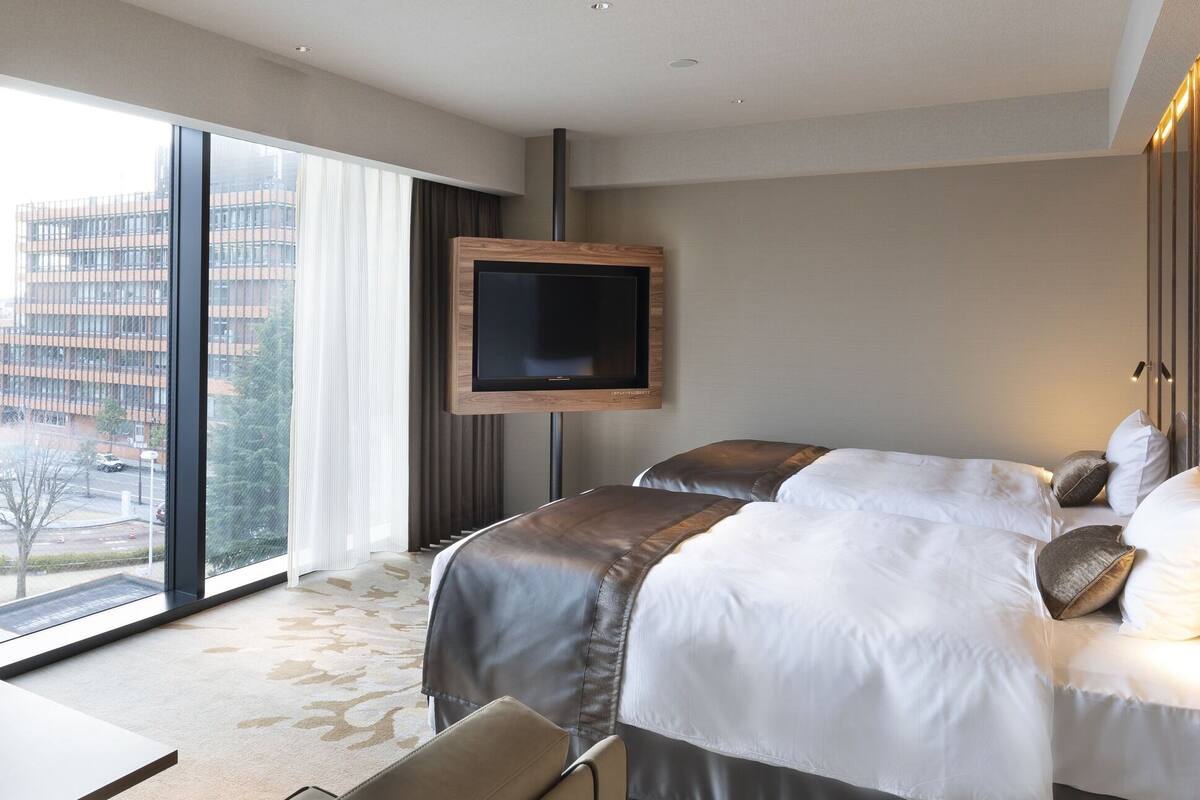 A hotel room with a large bed, a TV mounted on the wall, and a view of a cityscape through the window.