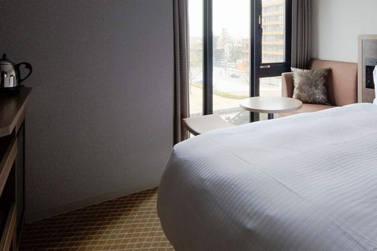 A hotel room with a large bed, a small round table, a desk with a kettle, and a view of the outside through a window.