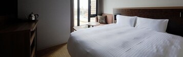 Habitación de hotel con una cama grande, una mesita redonda pequeña, un escritorio con una tetera y vistas al exterior a través de una ventana.