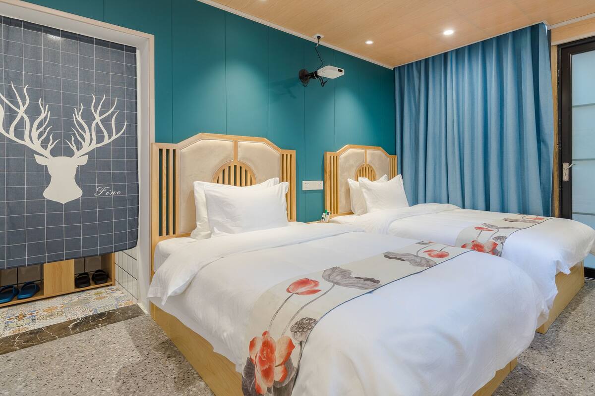 Ein Hotelzimmer mit zwei Betten, einem großen Fenster mit Vorhängen, einem Deckenventilator und einem Wandbild mit einem Hirschgeweih.