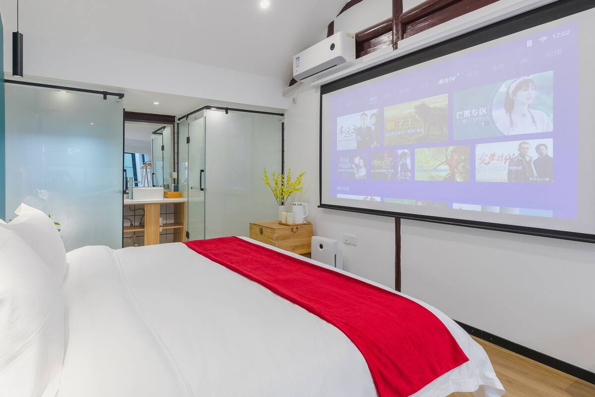 Ein Hotelzimmer mit einem großen Bett, einer roten Zierdecke, einem Nachttisch aus Holz und einem an der Wand montierten Fernseher, der Filmplakate zeigt.