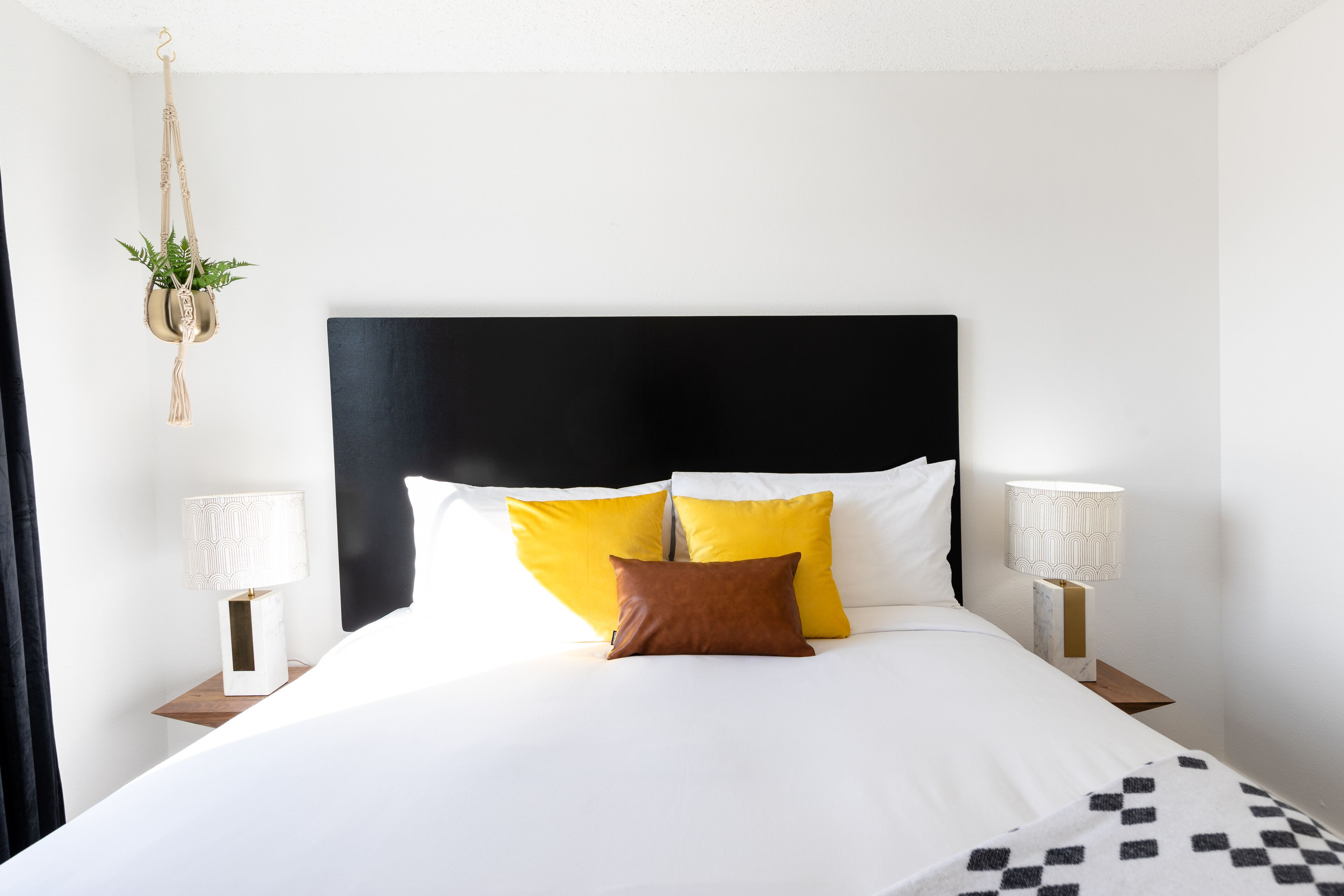 Una cama bien hecha con ropa de cama blanca y amarilla, dos lámparas de noche y una planta colgante.