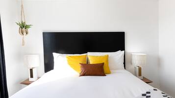 Una cama bien hecha con ropa de cama blanca y amarilla, dos lámparas de noche y una planta colgante.