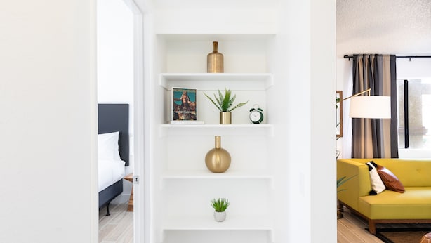 Un salon moderne avec une étagère blanche, un canapé jaune et un lit doté d’une tête de lit sombre.