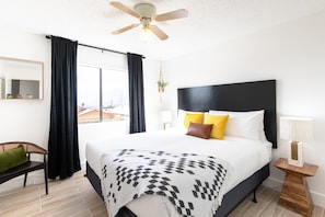 Una camera d'albergo moderna con un letto grande, una ventola a soffitto, una finestra con tende, un comodino con una lampada e una sedia.