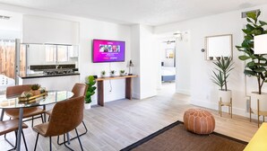 Un salon moderne avec une table à manger en verre, des fauteuils en cuir marron, un pouf rond et un téléviseur à écran plat fixé au mur.
