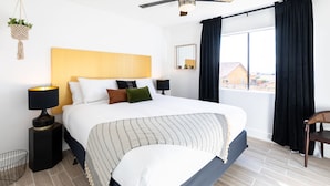 Une chambre d’hôtel avec un lit, un ventilateur de plafond, une fenêtre avec des rideaux, une table de chevet avec une lampe et une vue sur les bâtiments environnants.