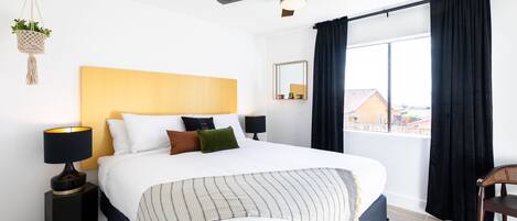 A hotel room with a bed, a ceiling fan, a window with curtains, a bedside table with a lamp, and a view of buildings outside.