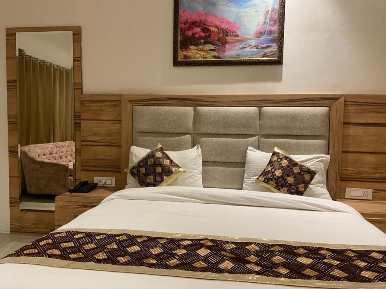 A hotel room with a large bed, a headboard with a padded design, two patterned pillows, a bedside table with a phone, and a painting above the bed.