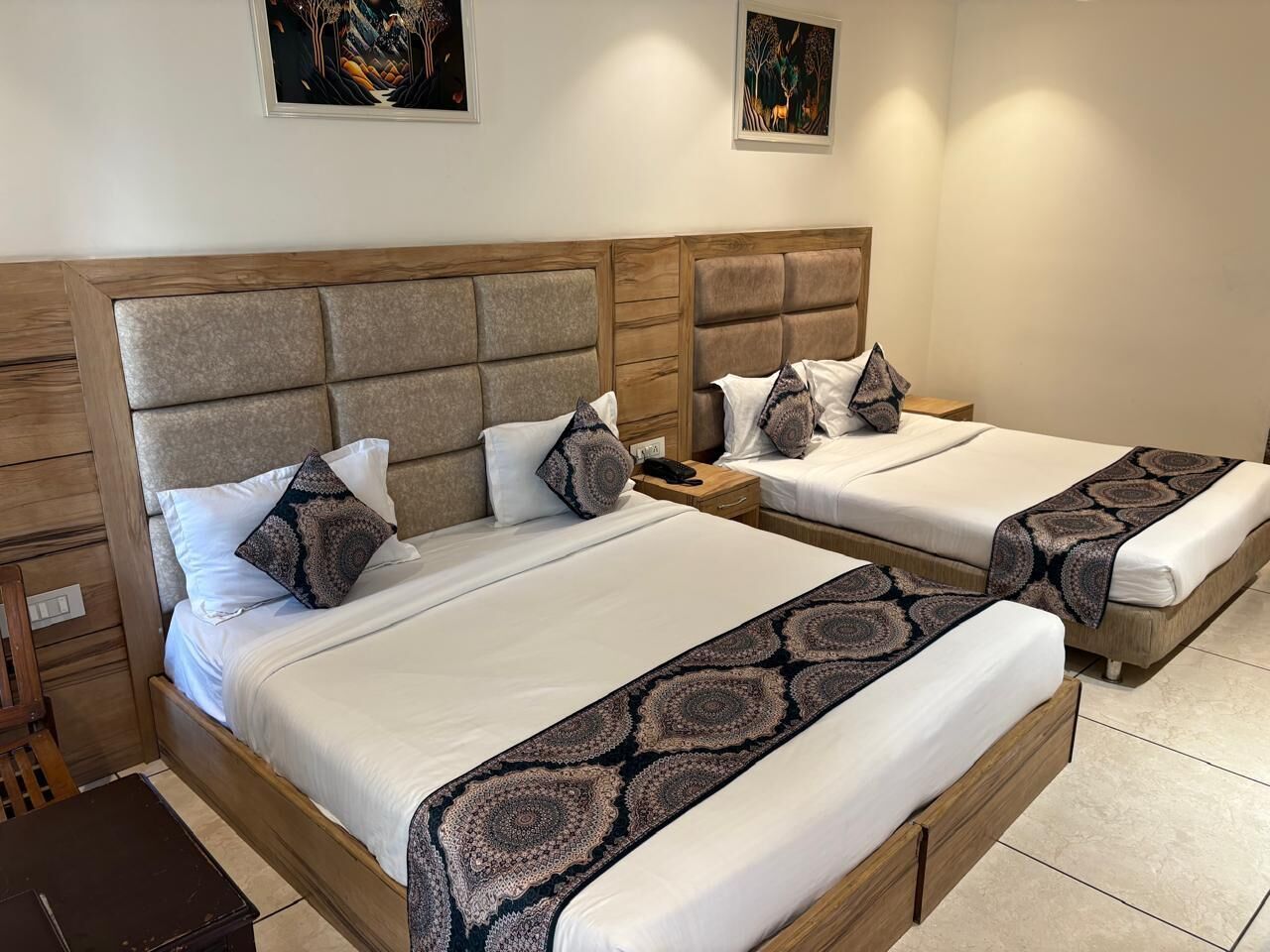 A hotel room with two beds, each with a wooden headboard and a patterned throw blanket. There are bedside tables with lamps and a small nightstand.