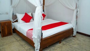Una cama con dosel, cubrelechos blancos y almohadas rojas, una mesita de noche de madera y piso de baldosas.