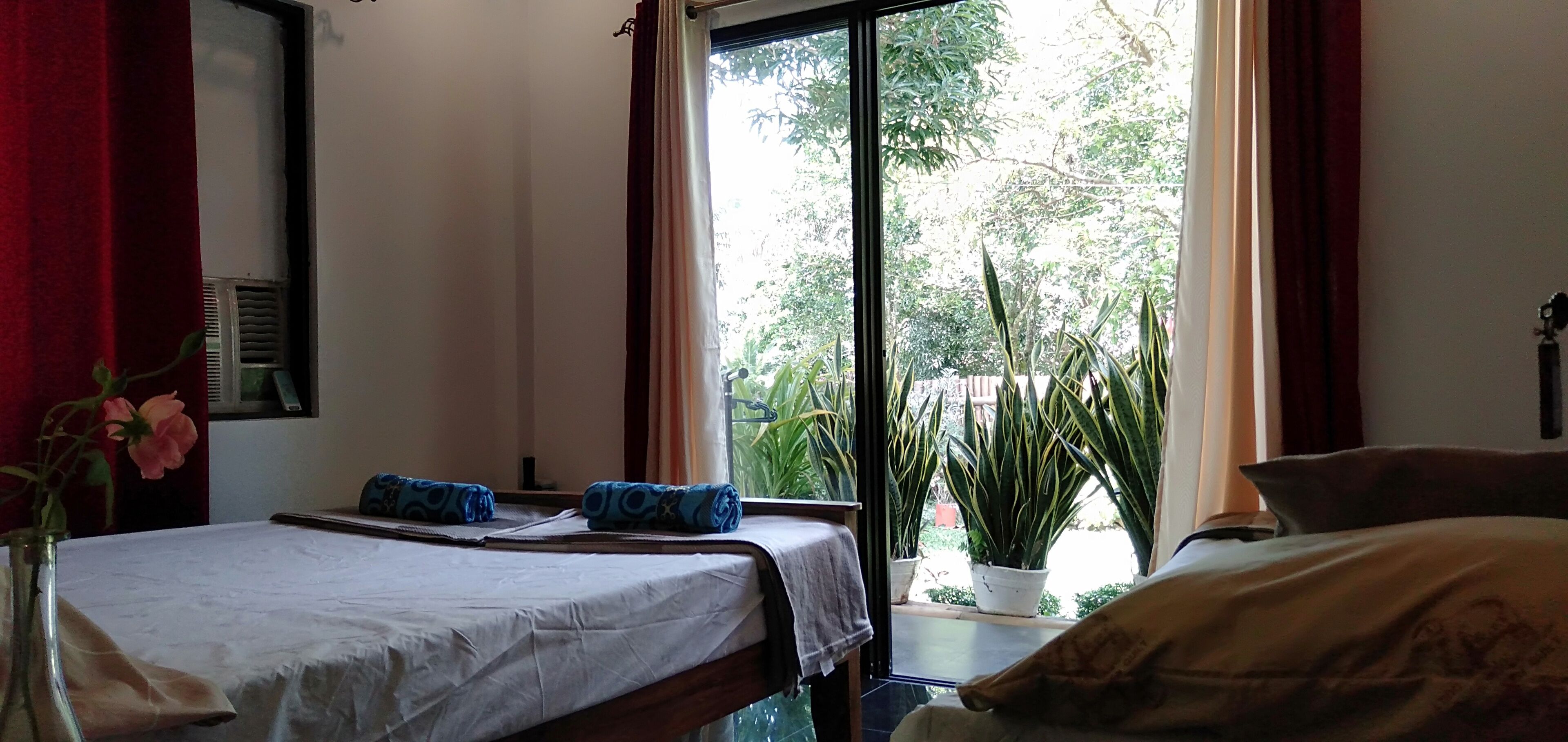 A hotel room with a bed, a small table, a vase with a flower, and a view of greenery outside.
