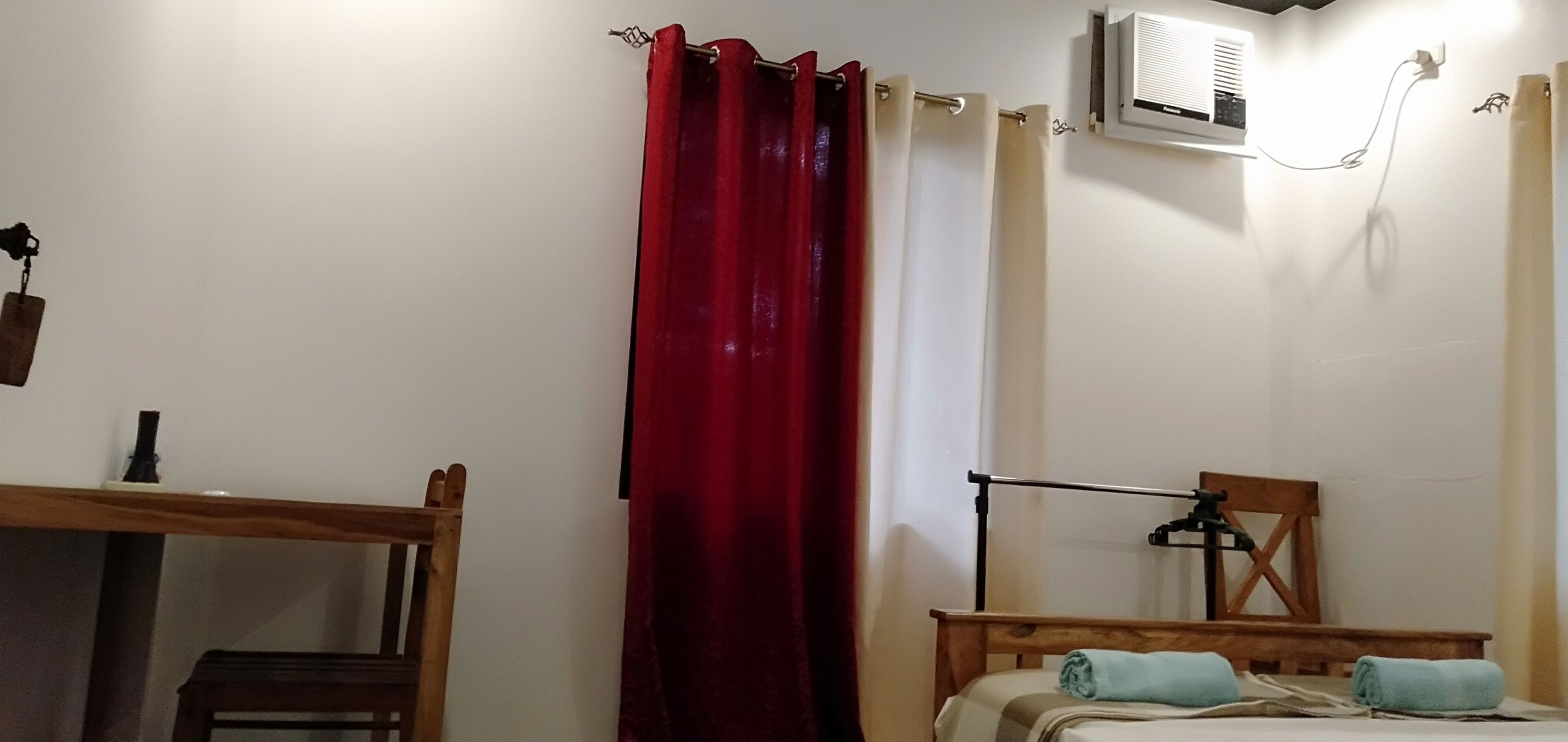 Una habitación con una cama, una silla de madera, un escritorio, una cortina roja y un equipo de aire acondicionado.