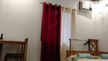 Una habitación con una cama, una silla de madera, un escritorio, una cortina roja y un equipo de aire acondicionado.