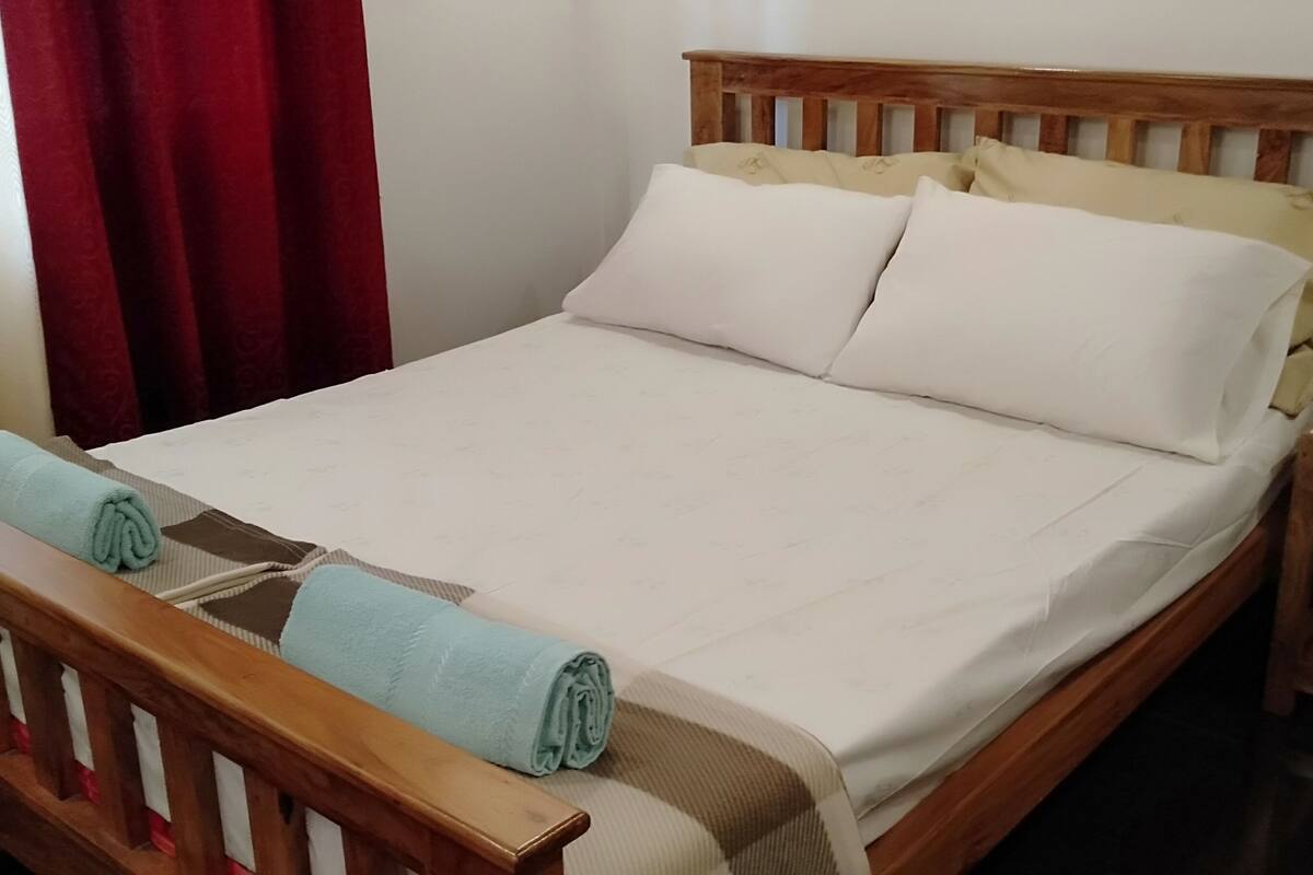 Una cama bien hecha con cabecera de madera, una mesita de noche pequeña y una cortina roja.