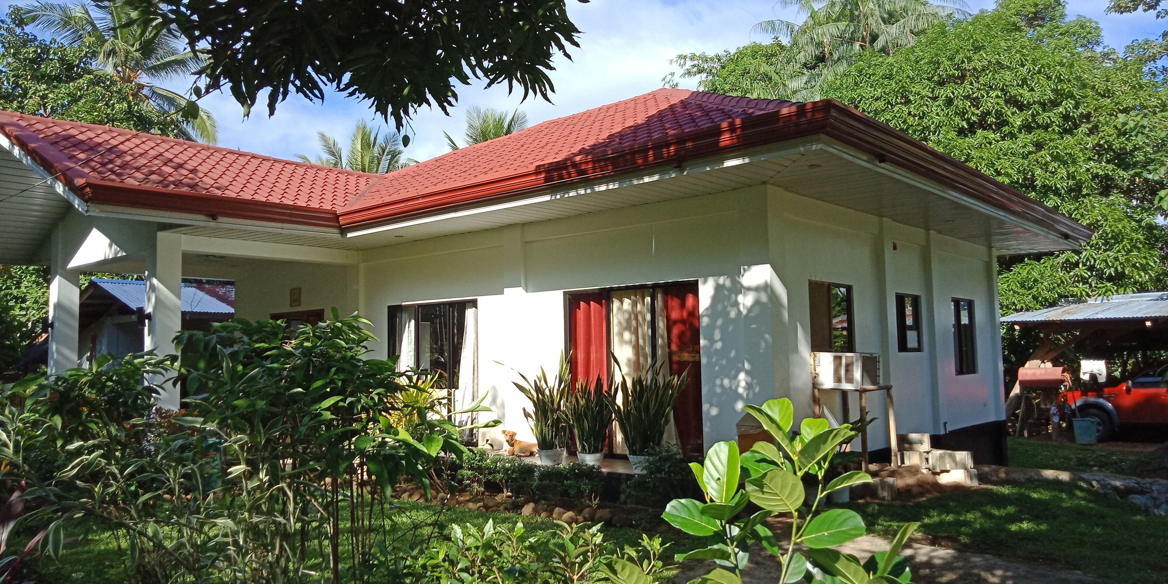 A single-story house with a red-tiled roof, a covered porch, and a small garden area with various plants.