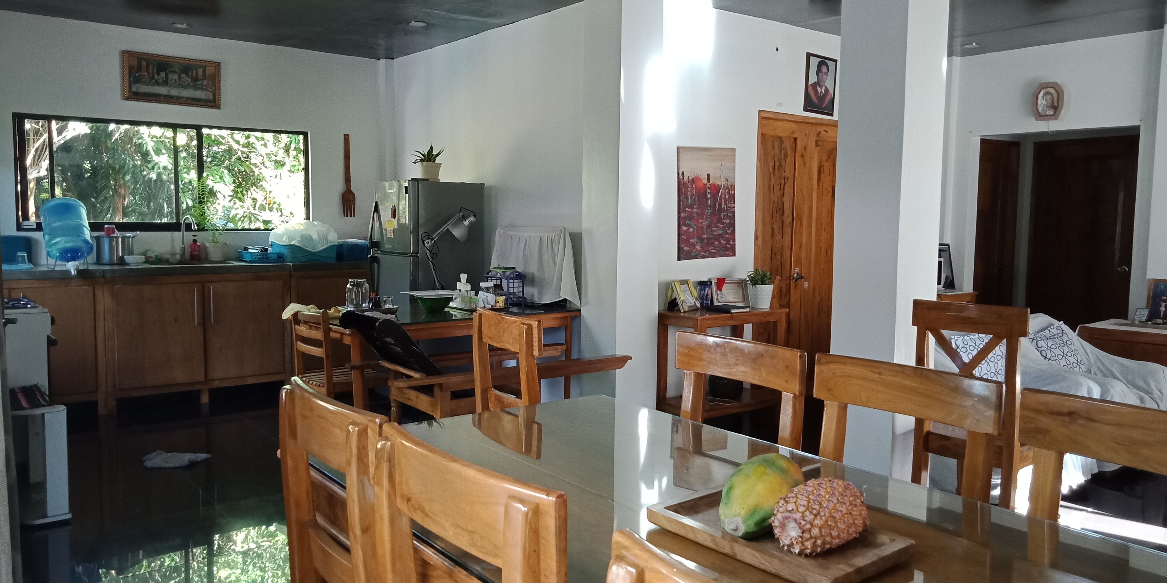 A modern kitchen with wooden cabinets, a dining table with chairs, a refrigerator, and a window with a view of greenery.