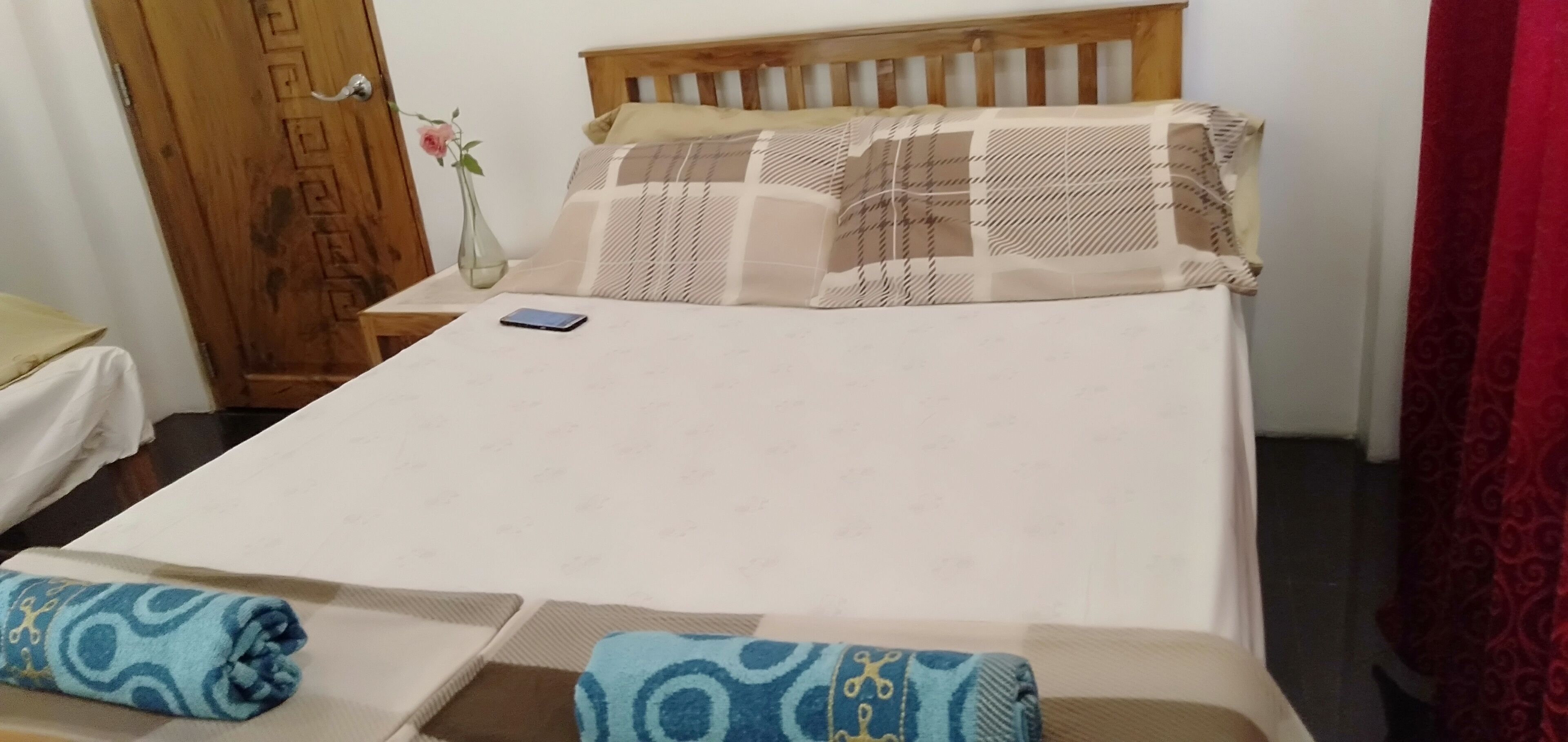 Un lit bien fait, avec un téléphone intelligent posé dessus, une tête de lit en bois et un vase avec une fleur sur la table de chevet.