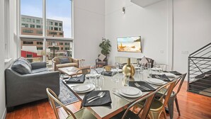 Un'area pranzo moderna con un tavolo in vetro apparecchiato per sei persone, circondato da sedie in legno. Un'ampia finestra offre vista sulla città.