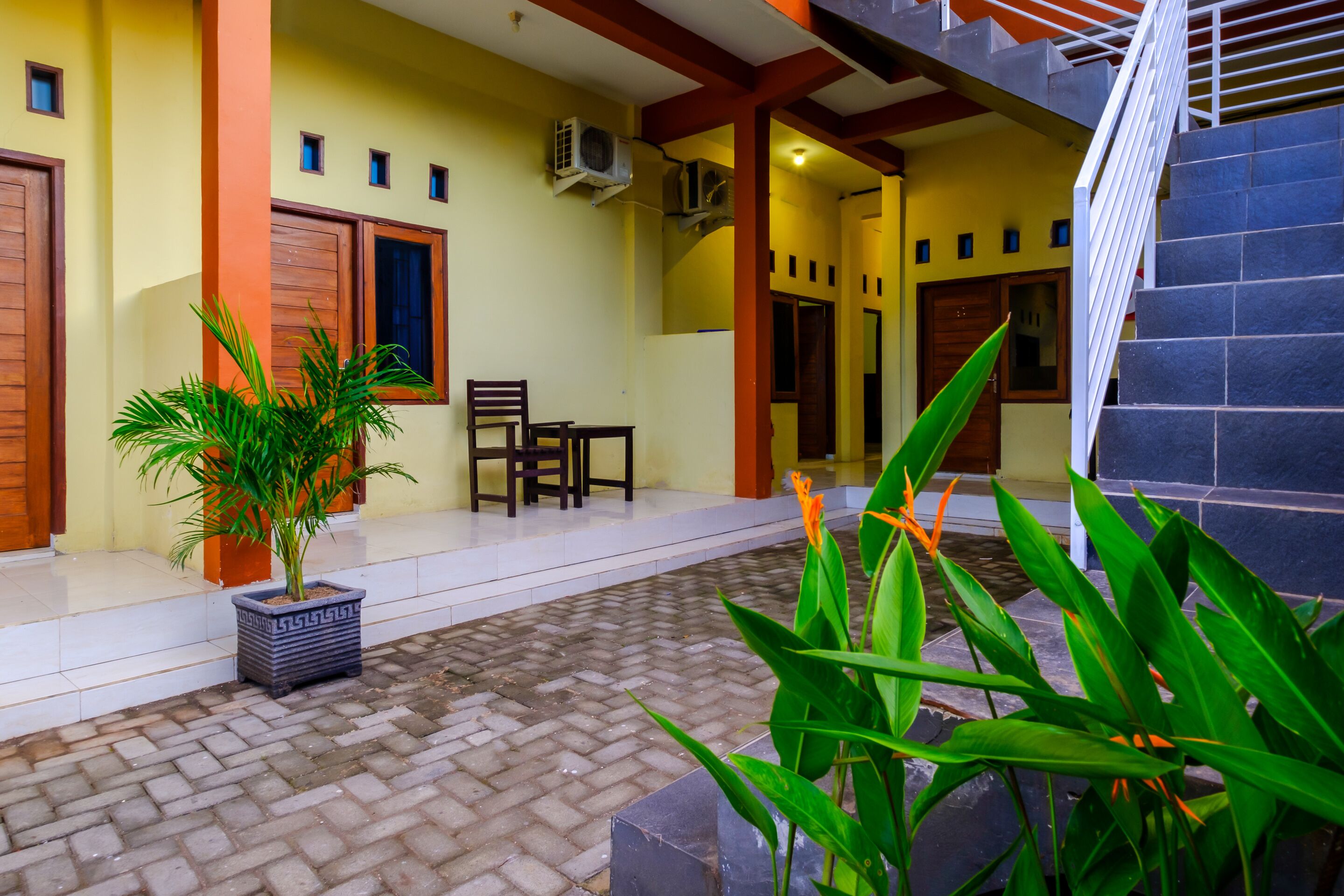 A covered outdoor area with a tiled floor, a potted plant, a wooden chair, and a staircase.