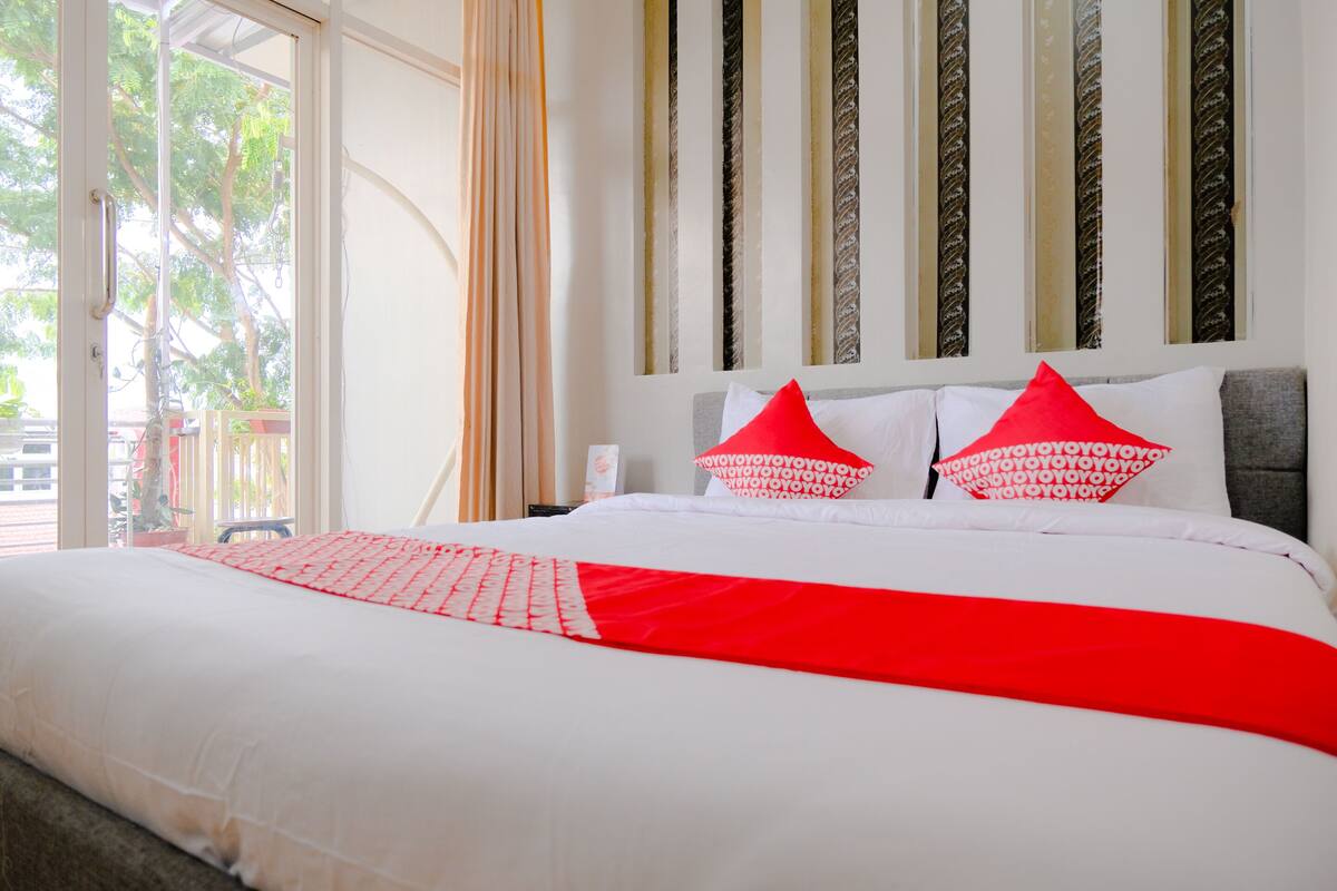 A neatly made bed with white and red bedding, a grey headboard, and a view of the outdoors through a glass door.