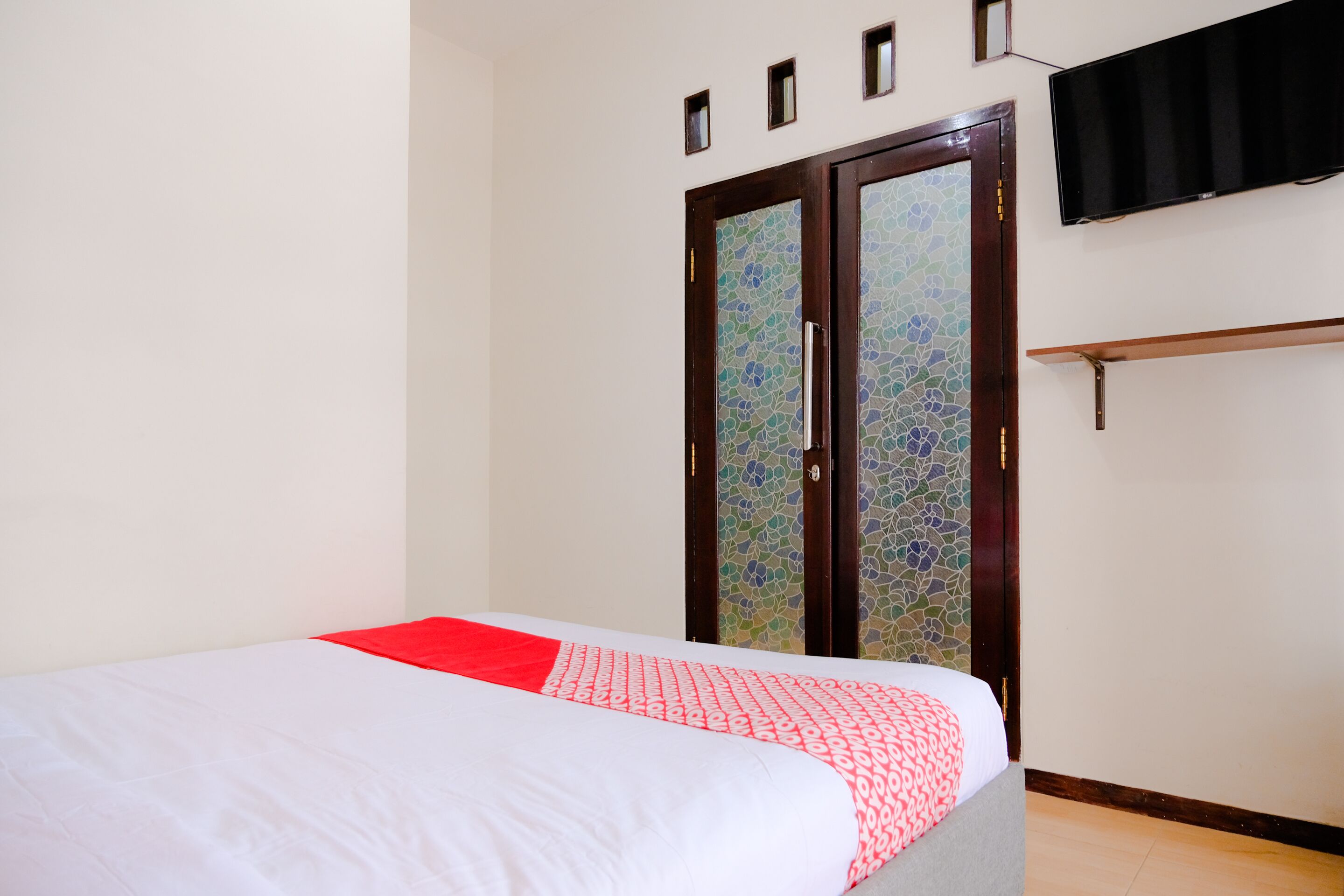 Une chambre à coucher avec un lit, un coussin décoratif rouge, une armoire dont la porte est ornée de motifs en mosaïque et un téléviseur fixé au mur.