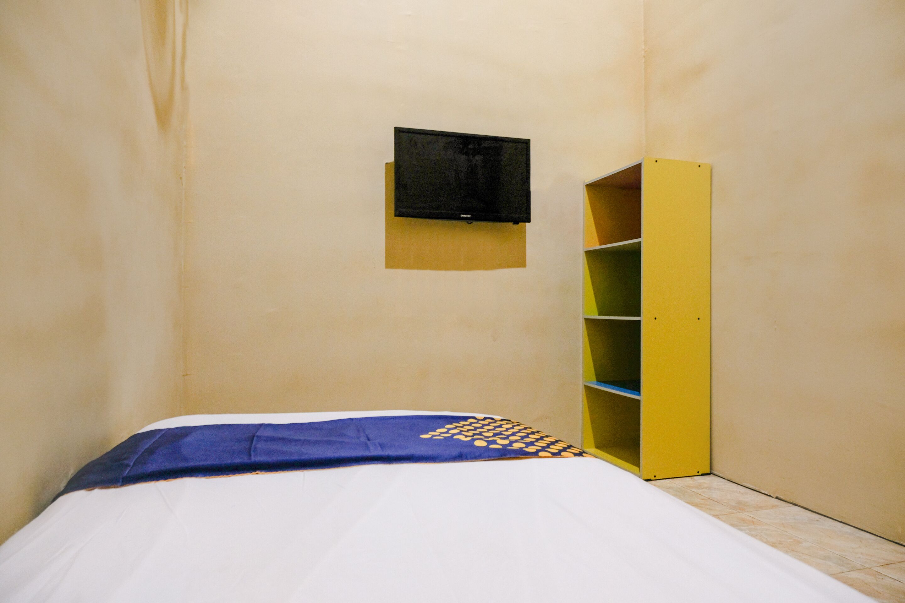 Une petite pièce étroite, avec un lit, un téléviseur fixé au mur et une étagère jaune.