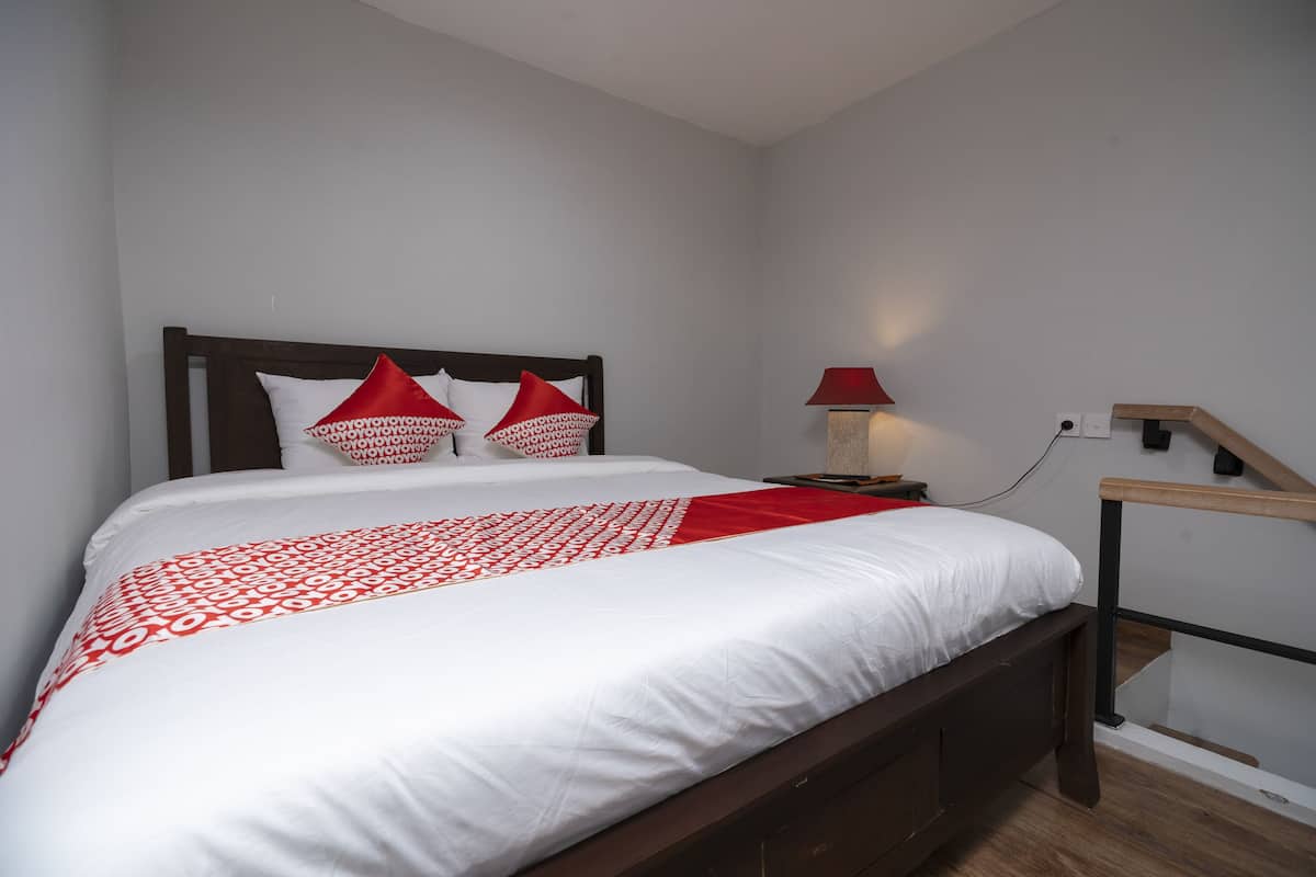 Ein ordentlich gemachtes Bett mit einer Bettdecke in Weiß und Rot, ein Holz-Kopfteil und zwei rote Kissen. Ein Nachttisch mit roter Lampe und Steckdose steht daneben. Am Fußende des Bettes befindet sich ein Holzstuhl.