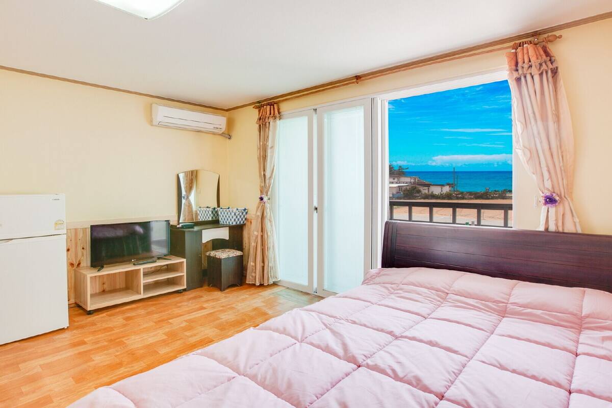 A bedroom with a bed, a TV, a small fridge, a desk, and a window with a view of the sea.