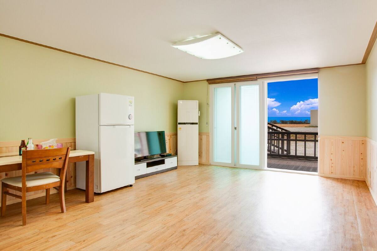 A spacious room with a wooden floor, a kitchenette with a refrigerator, a TV, and a dining area with a wooden table and chair.