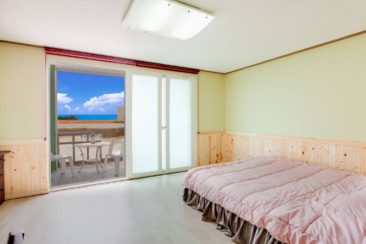 A bedroom with a bed, a sliding glass door leading to a balcony with a view of the beach and ocean.