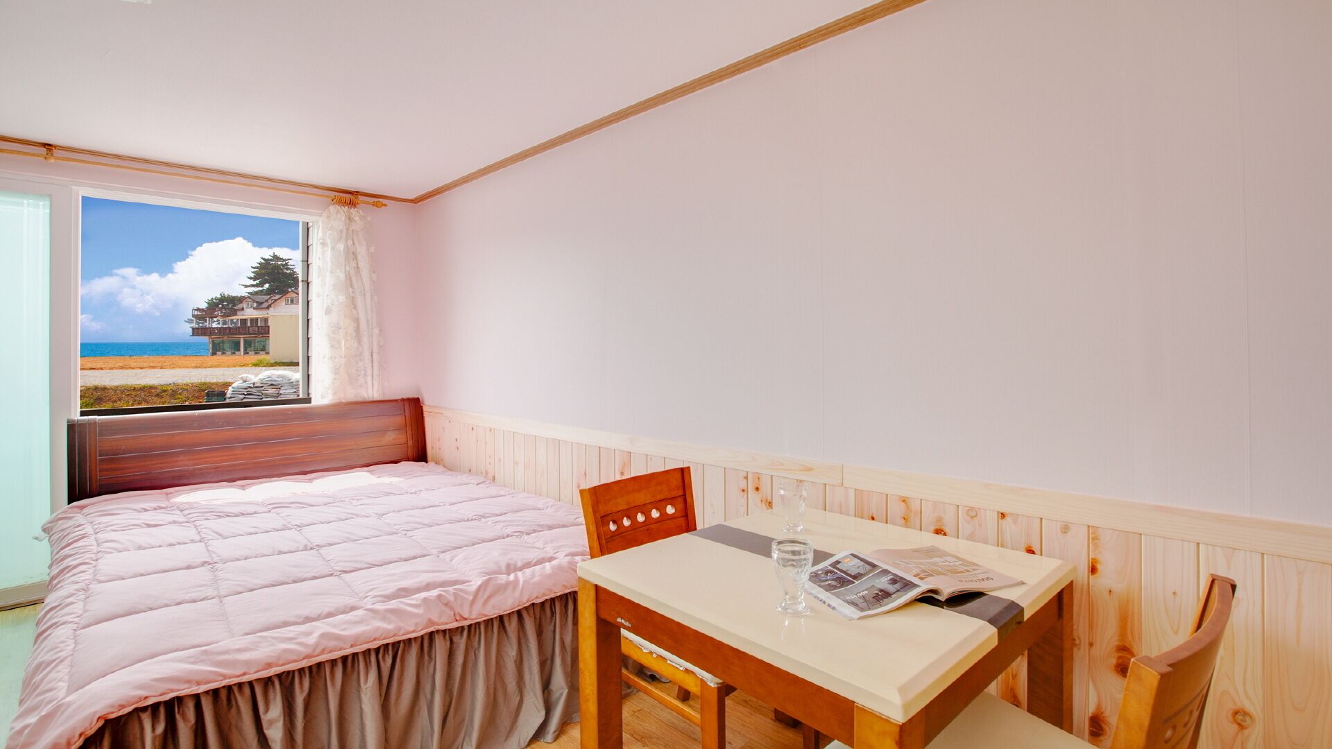 Um quarto com uma cama, uma mesa com um copo e uma revista, e uma janela com vista para uma praia e uma casa.