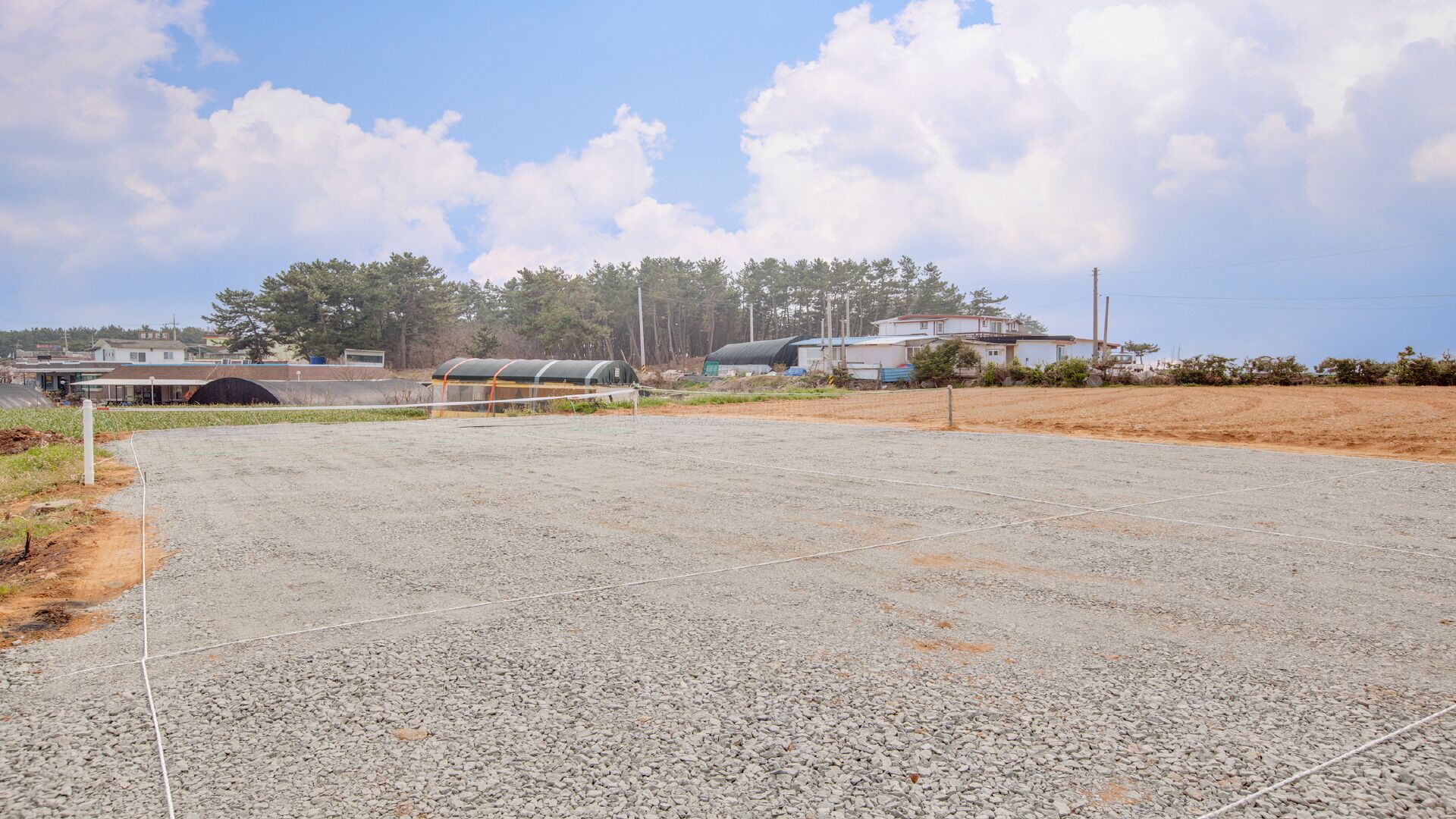 A gravel parking area with a sports court, surrounded by buildings and trees.
