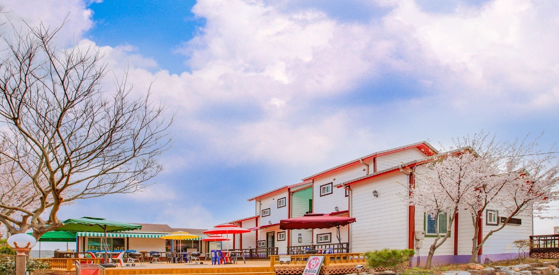 A building with a red roof and white walls, a patio area with tables and chairs, and a large tree with bare branches. - Vellise Pension (Taean)