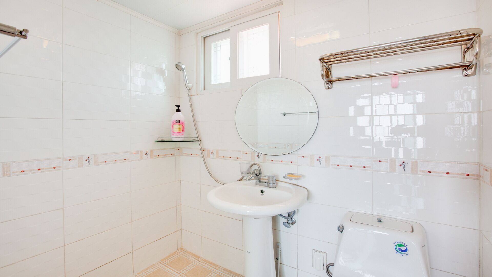 A bathroom with white tiles, a round mirror, a sink, a shower, and a window.