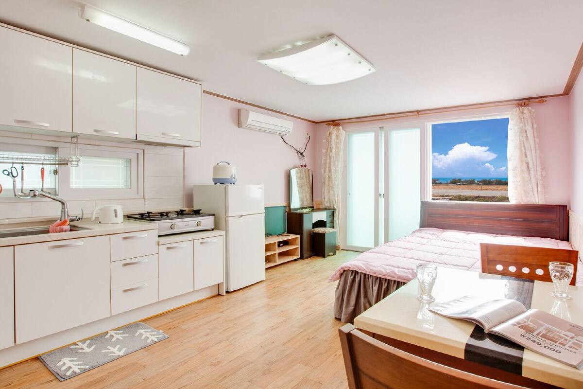 A compact room with a kitchenette, a bed with a view, and a small table with a magazine.