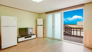 A room with a wooden floor, a white refrigerator, a flat-screen TV, and a sliding glass door leading to an outdoor deck with a view of the beach. - Vellise Pension (Taean)