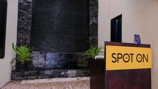 A reception area with a 'SPOT ON' sign, a black textured wall, and two potted plants. - SPOT ON 2431 Tf Homestay (Caruban)