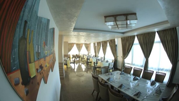 A dining area with a long table set for a meal, surrounded by chairs. There are multiple windows with curtains, and a ceiling light fixture is visible. - Elgel Hotel and Spa (Addis Ababa)