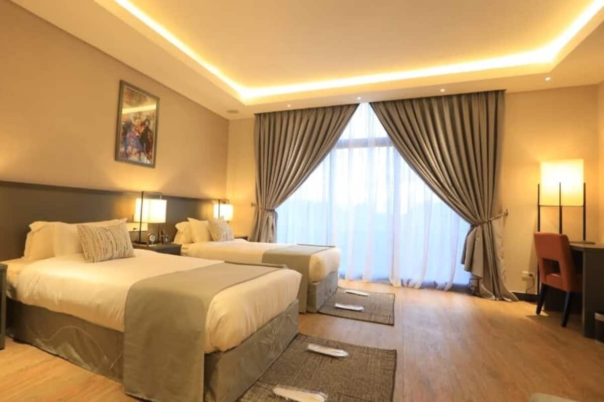A hotel room with two beds, a desk, a chair, and a large window with curtains.