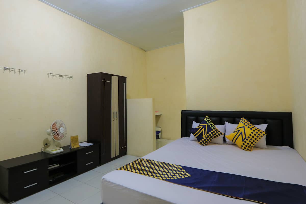 A hotel room with a bed, a wardrobe, a desk with a fan, and a small table with a lamp.