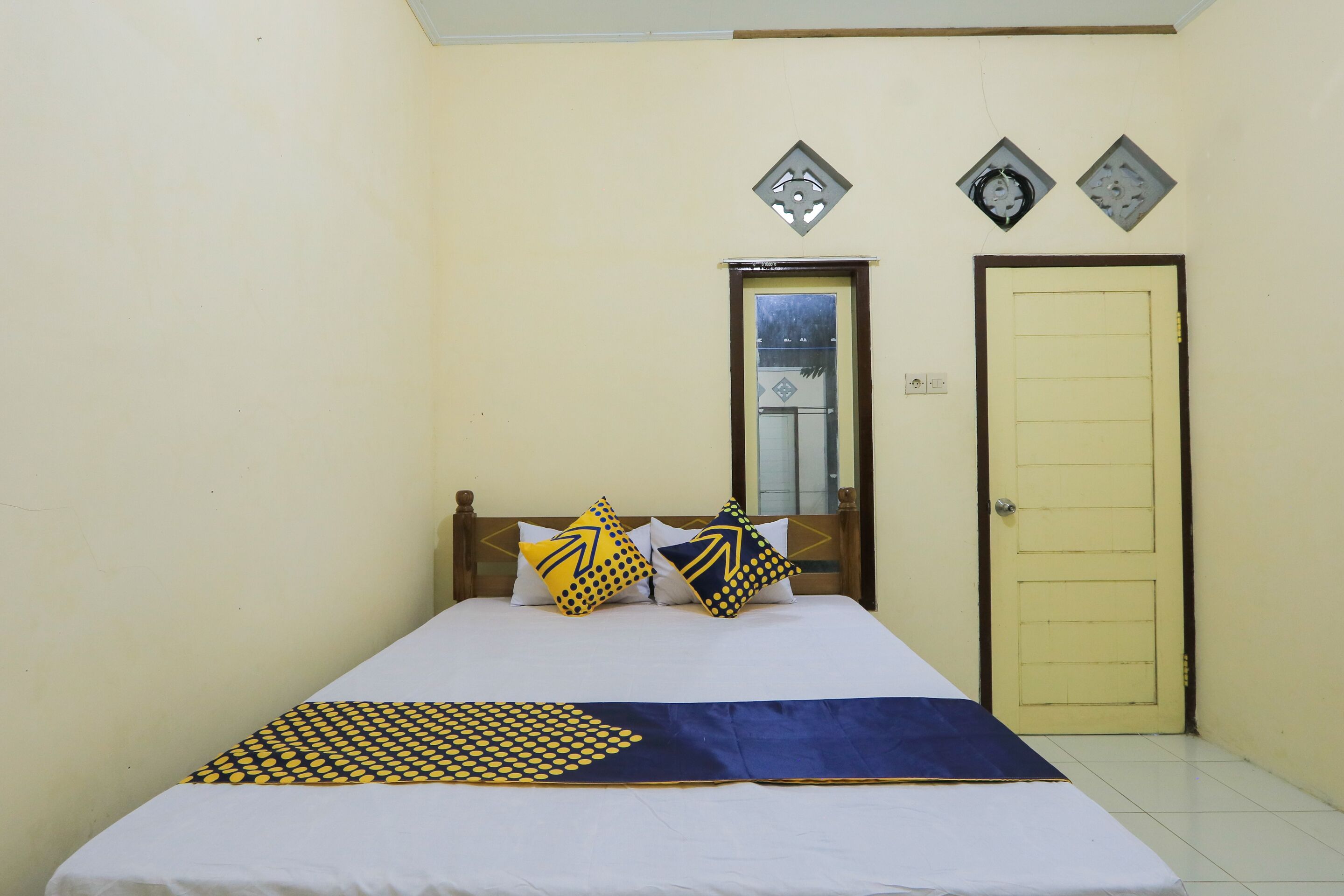 A single bed with a patterned bedspread, two pillows with arrow designs, a wooden headboard, and a plain door.
