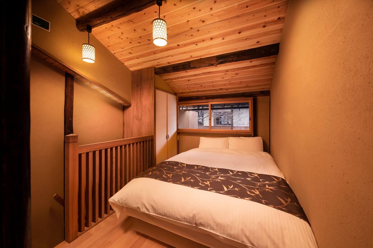 Une chambre avec un plafond en bois, un lit recouvert d’une couvre-lit à motifs et une fenêtre donnant sur des arbres.