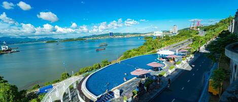 A waterfront resort with a swimming pool, a road with parked cars, and a view of the river and distant buildings.