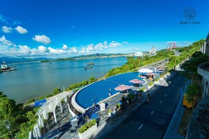 Un resort frente al agua con piscina, una calle con coches estacionados y vistas al río y edificios al fondo.