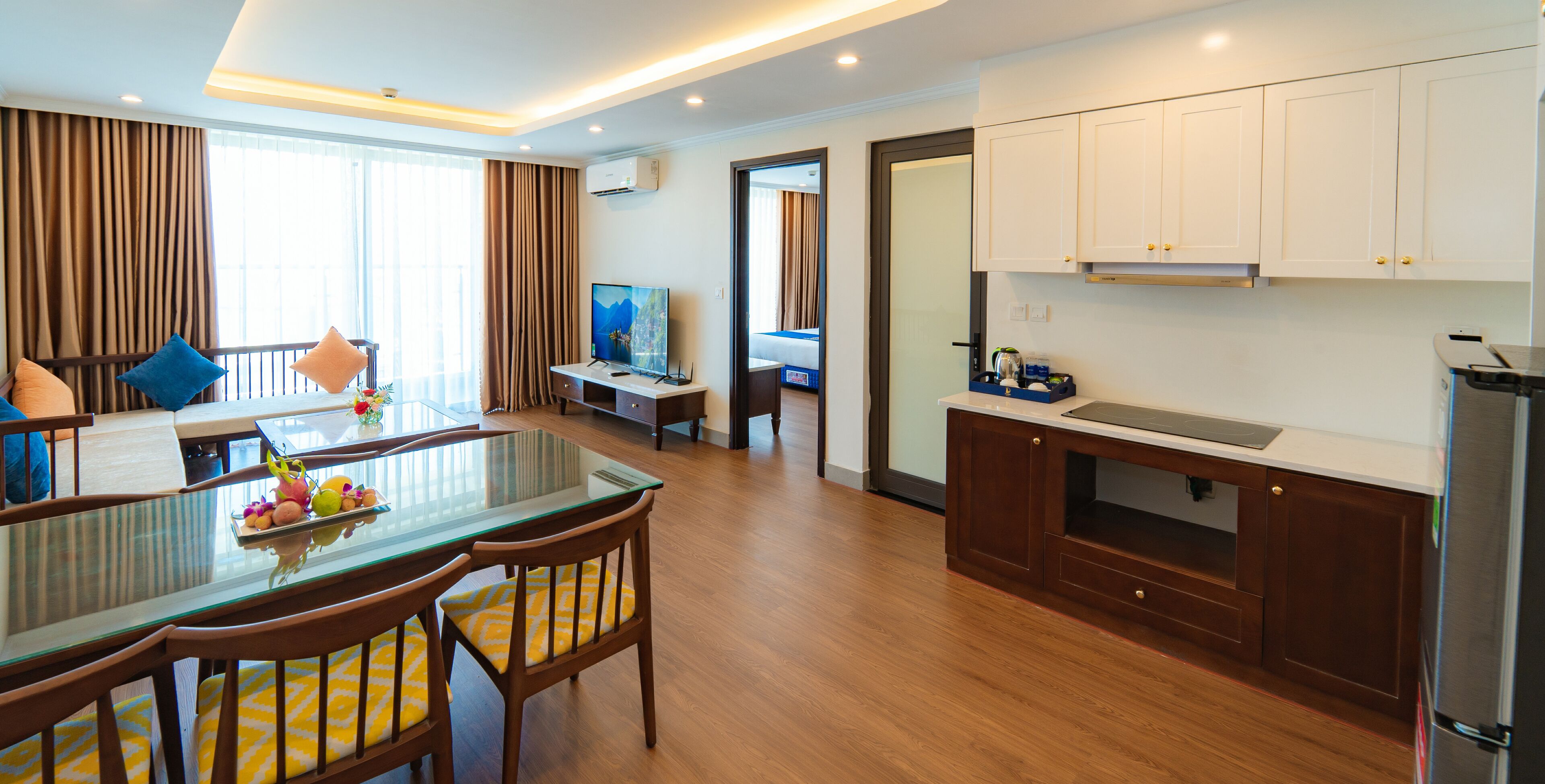 A modern hotel room with a dining area, kitchen, and a large window with curtains.