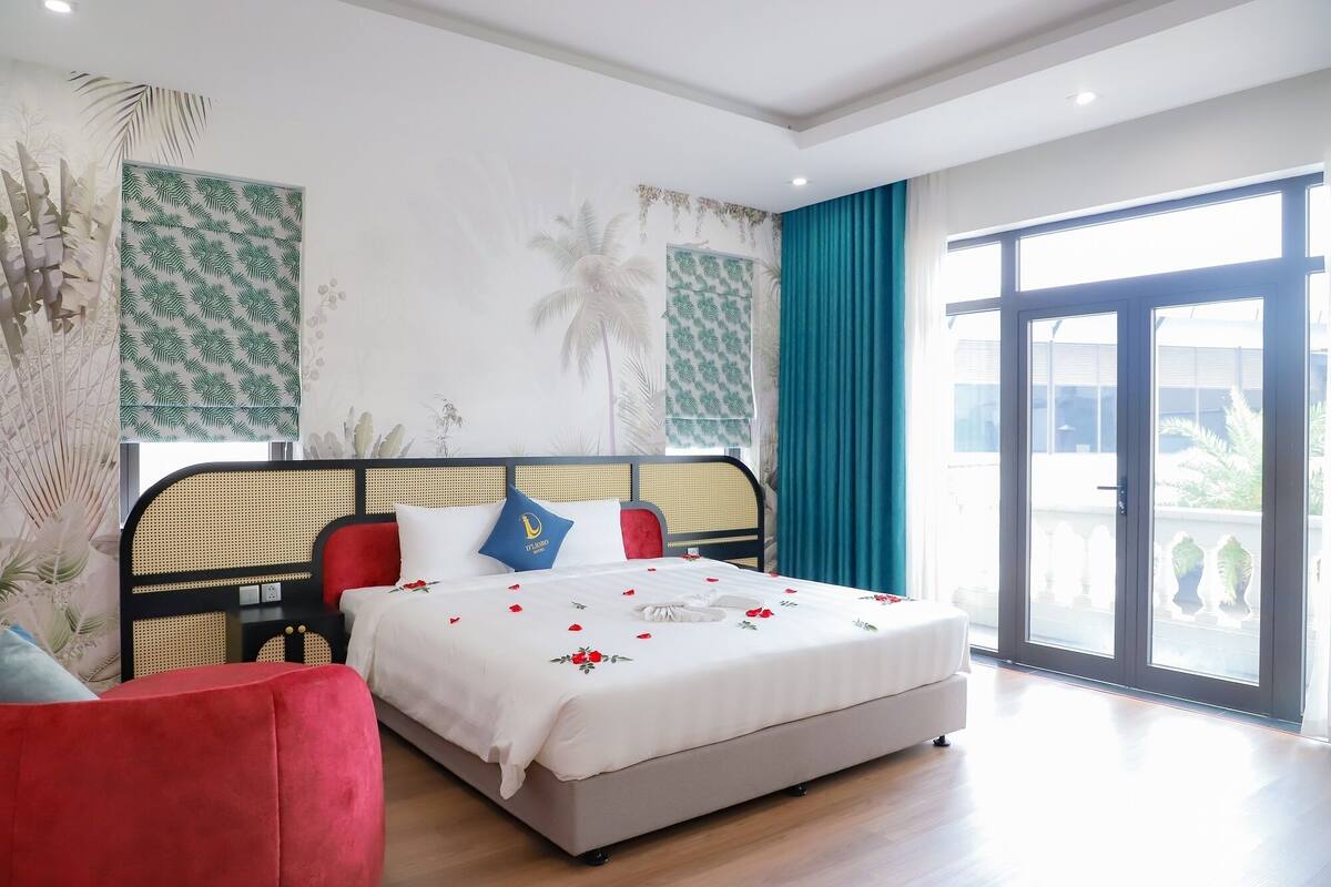 Une chambre d’hôtel avec un lit, un fauteuil rouge, une grande fenêtre donnant sur un beau paysage et un balcon.