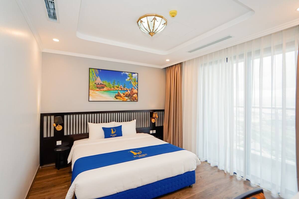 Une chambre d’hôtel avec un grand lit, un tableau représentant une scène de plage, un lustre et une vue sur l’extérieur à travers des rideaux transparents.