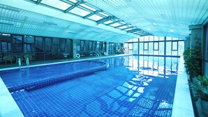 An indoor swimming pool with clear glass ceilings, a tiled floor, and a view of the city through large windows.