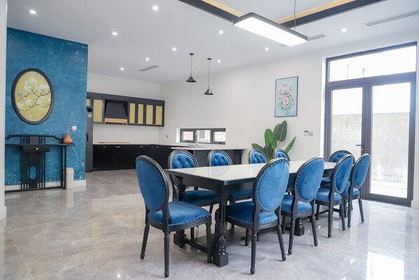 A modern dining area with a large table and blue upholstered chairs, a kitchen area with black cabinets, and a large window providing natural light.